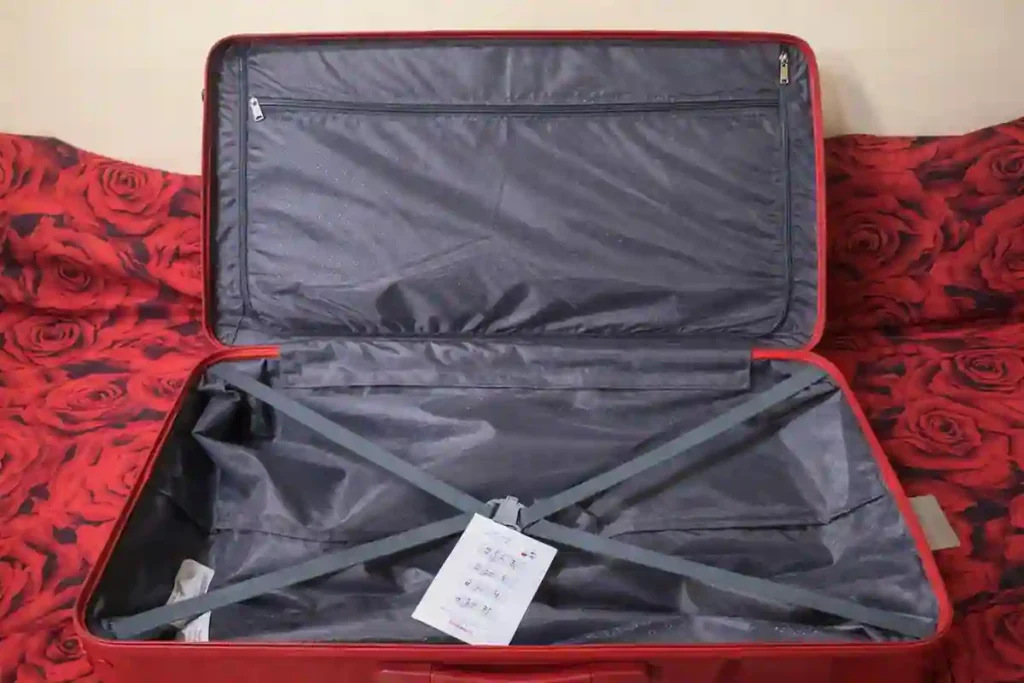 The interior of an American Tourister hard-shell trolley bag opened flat, featuring a full-length zippered divider, smooth fabric lining, and adjustable cross compression straps to keep clothes secure